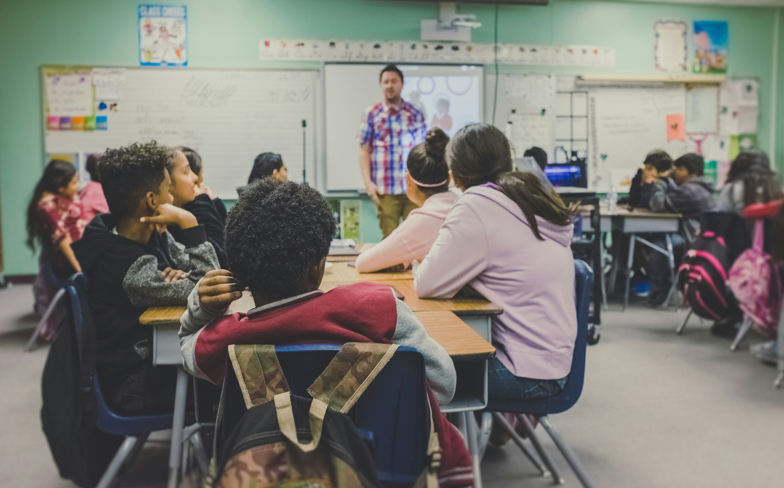 Classroom with students seated at desks, facing a teacher standing at the front, near a whiteboard and projector screen.
