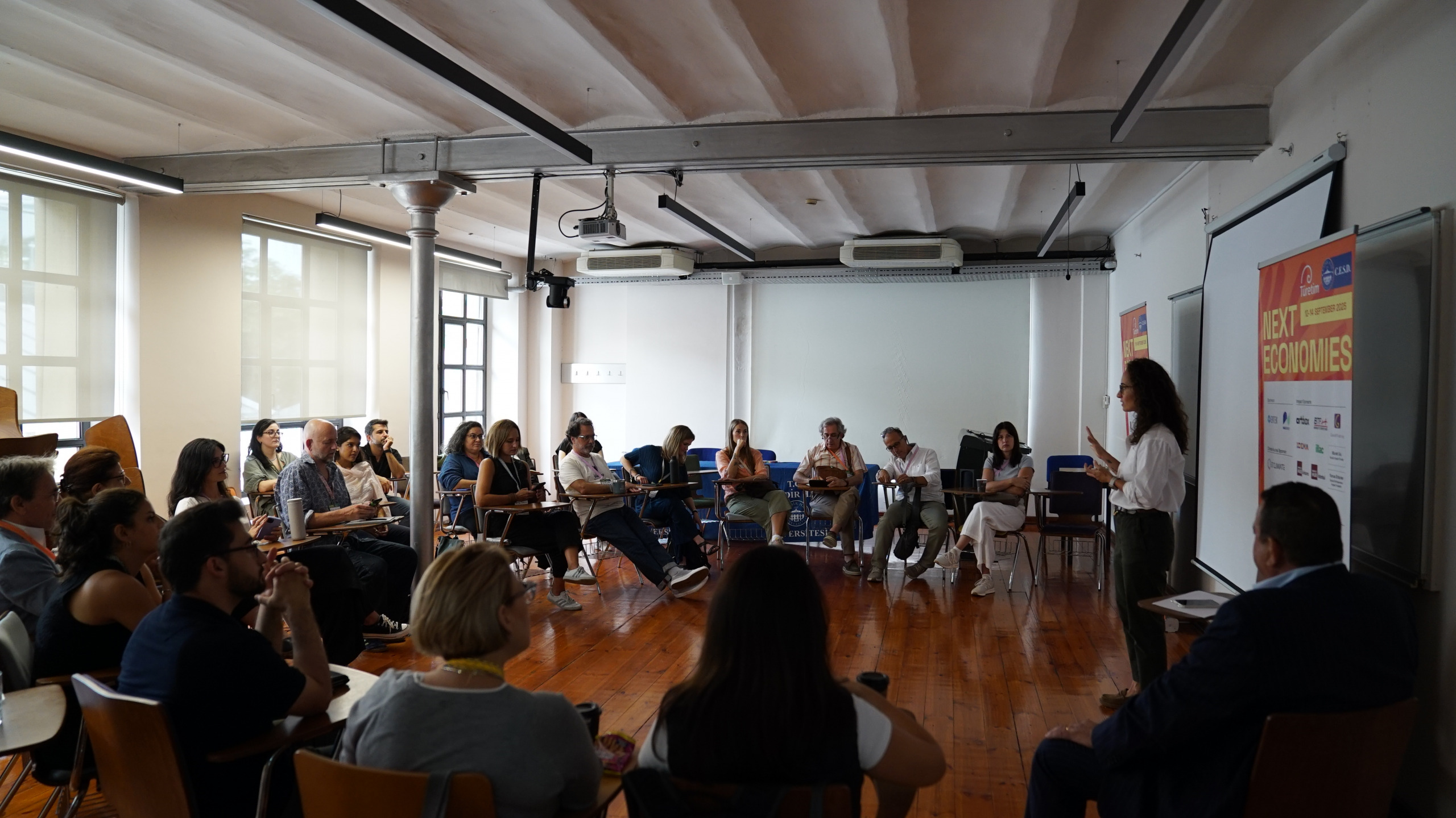 Individuals seated in a classroom during a presentation. A speaker stands in front of a screen displaying a banner titled Next Economies. The room has wooden floors, a ceiling with exposed beams, and large windows.