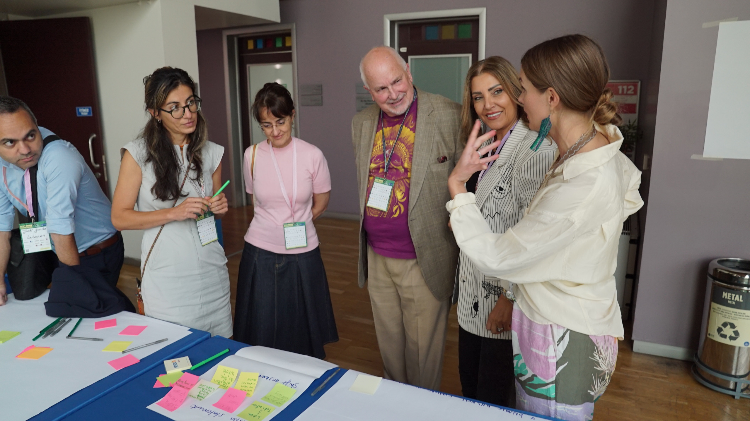 A group of six people standing together indoors, engaging in discussion. They are gathered around a table with paper and colorful sticky notes. The setting appears to be a casual meeting or workshop.