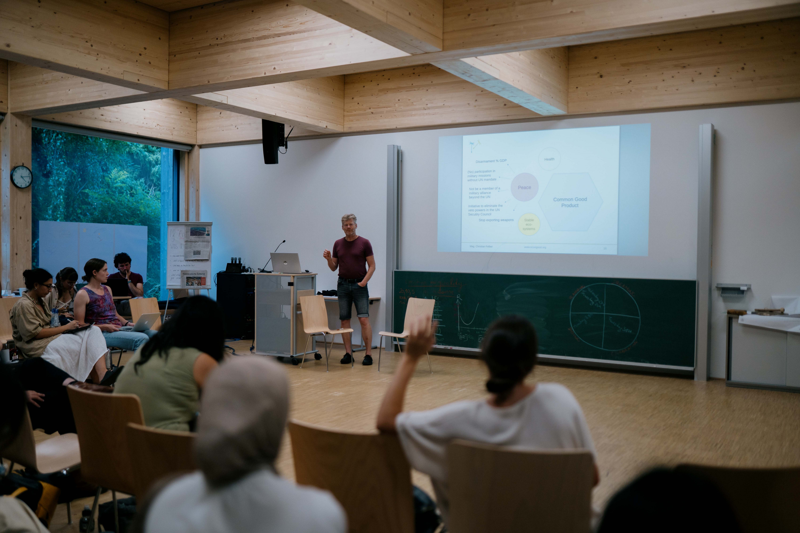 A person stands in front of a group giving a presentation in a bright classroom. A slide with a diagram is projected on the wall, and there is a chalkboard with graphs behind the speaker. The audience is seated and appears engaged.