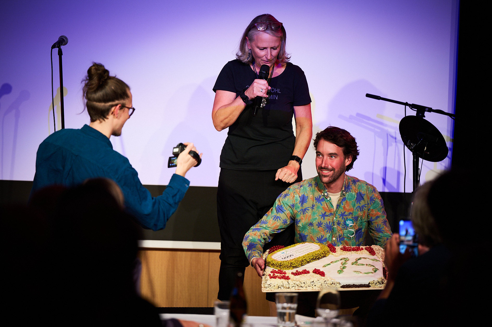 Person sitting on stage holding a large decorated cake with the number 75, while another person stands nearby speaking into a microphone. An individual in the foreground photographs the scene.