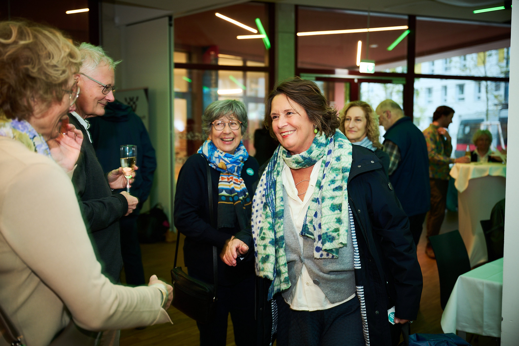 Group of people smiling and conversing at an indoor event, with one person holding a drink. The setting includes tables and coats visible in the background.